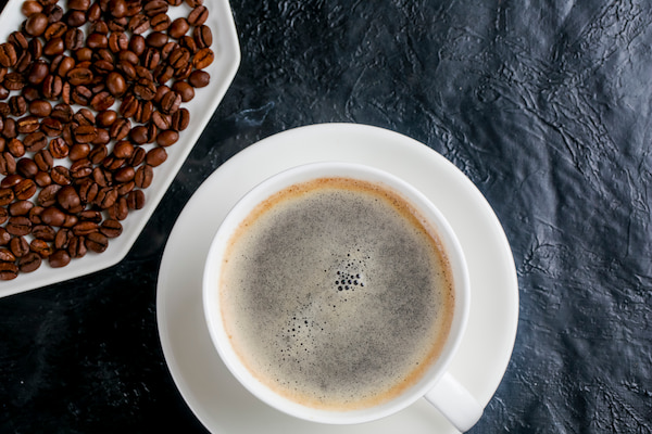 une tasse blanche de café noir sur une soucoupe est posée à côté d'une assiette remplie de grains de café torréfiés sur une surface à la texture sombre.