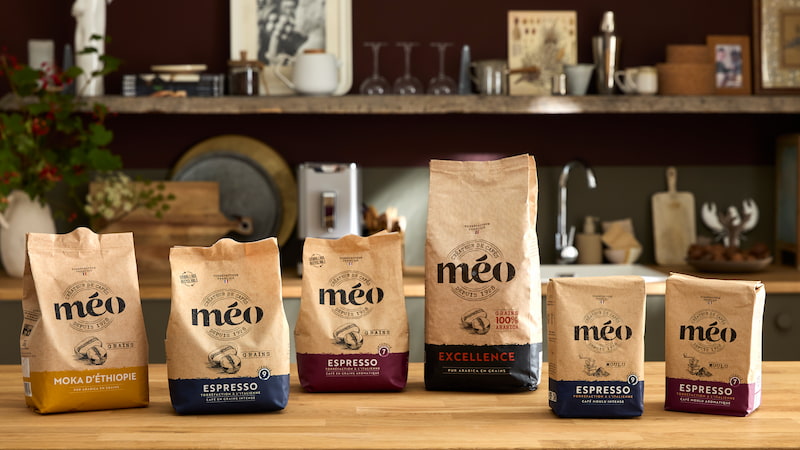 six sacs de café méo de tailles et de mélanges différents sont disposés en ligne sur un comptoir en bois, sur fond de cuisine avec des étagères, des tasses et des éléments de décoration.