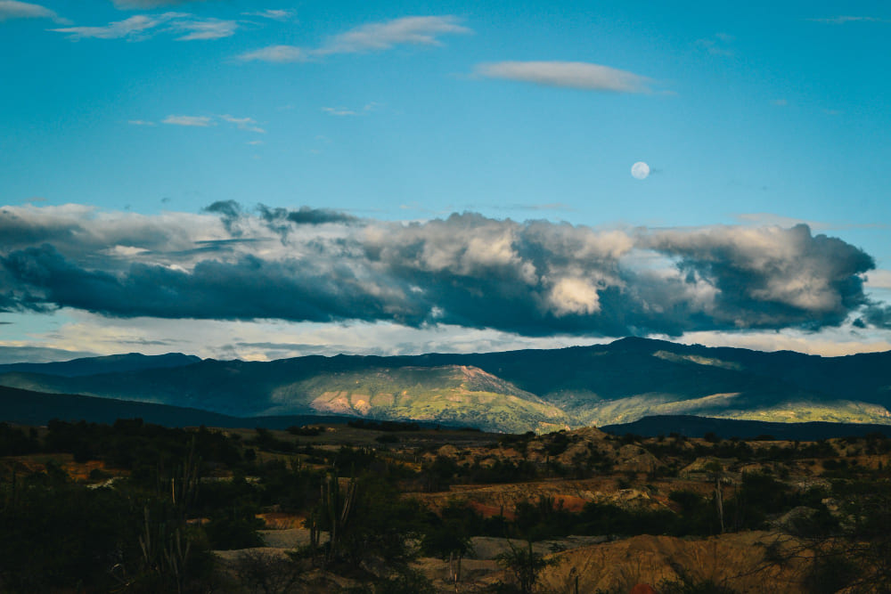 un paysage de montagnes sous un ciel bleu, des nuages épars, une lune visible et la lumière du soleil illuminant une partie des collines.